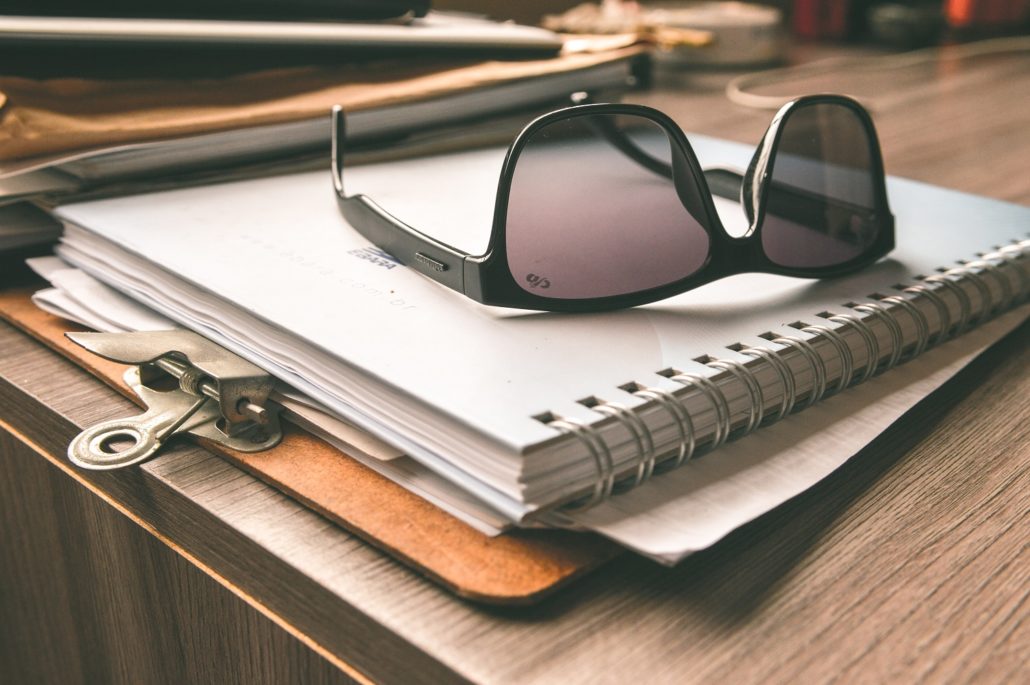 clipboard on brown wooden table with a stack of appraisal paperwork and sunglasses on top of it.