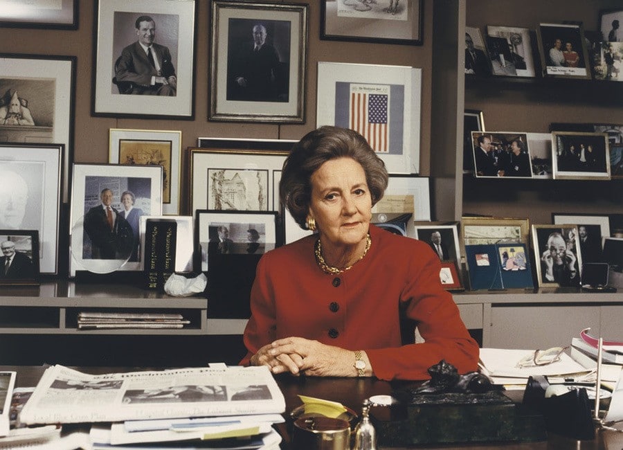 Katharine Graham in a red suit sitting at her desk in her office surrounded by photos.