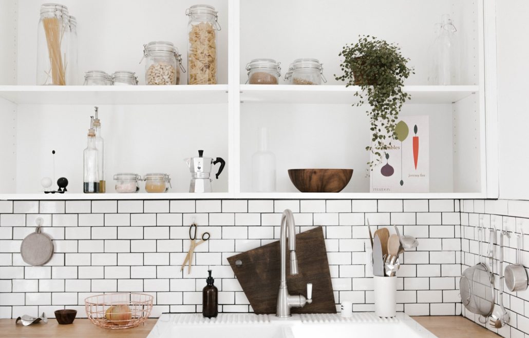 Photo of a white kitchen sink with subway tile back splash and open shelving. 