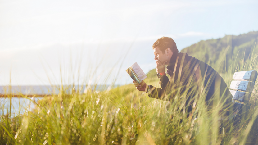 Man sitting on bench in a field reading a book ocean view