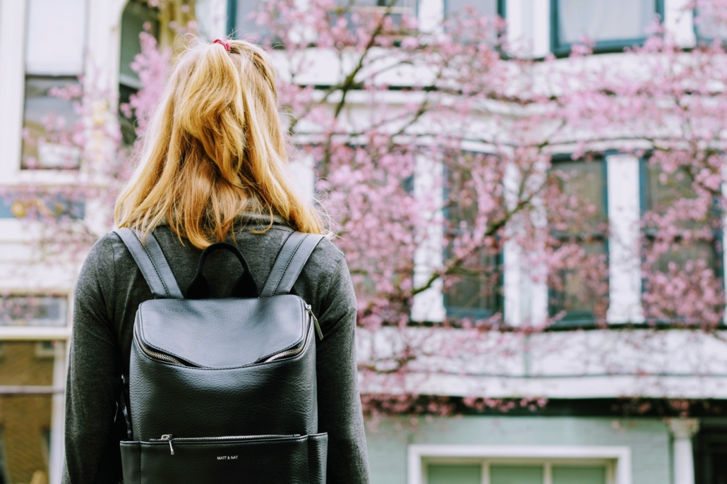 blonde Texas woman facing away from the camera wearing a black backpack looks at pink blossoming trees and local houses for sale
