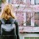blonde Texas woman facing away from the camera wearing a black backpack looks at pink blossoming trees and local houses for sale