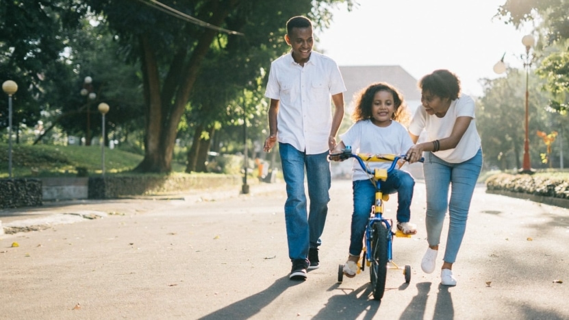 a millennial mother and father wearing white shirts teach their daughter to ride a bicycle outside in front of their Texas home