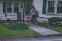 woman and young son carrying a red backpack walk toward their white house with green grass and yellow flowers in the front yard