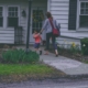 woman and young son carrying a red backpack walk toward their white house with green grass and yellow flowers in the front yard