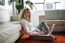 young woman with curly hair sitting on living room floor looking at mortgage rates on a laptop