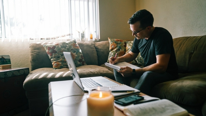 Man sits on green living couch while researching loan forbearance with a laptop and notepad. Most loan servicers recoup loan payments at the end of forbearance periods.