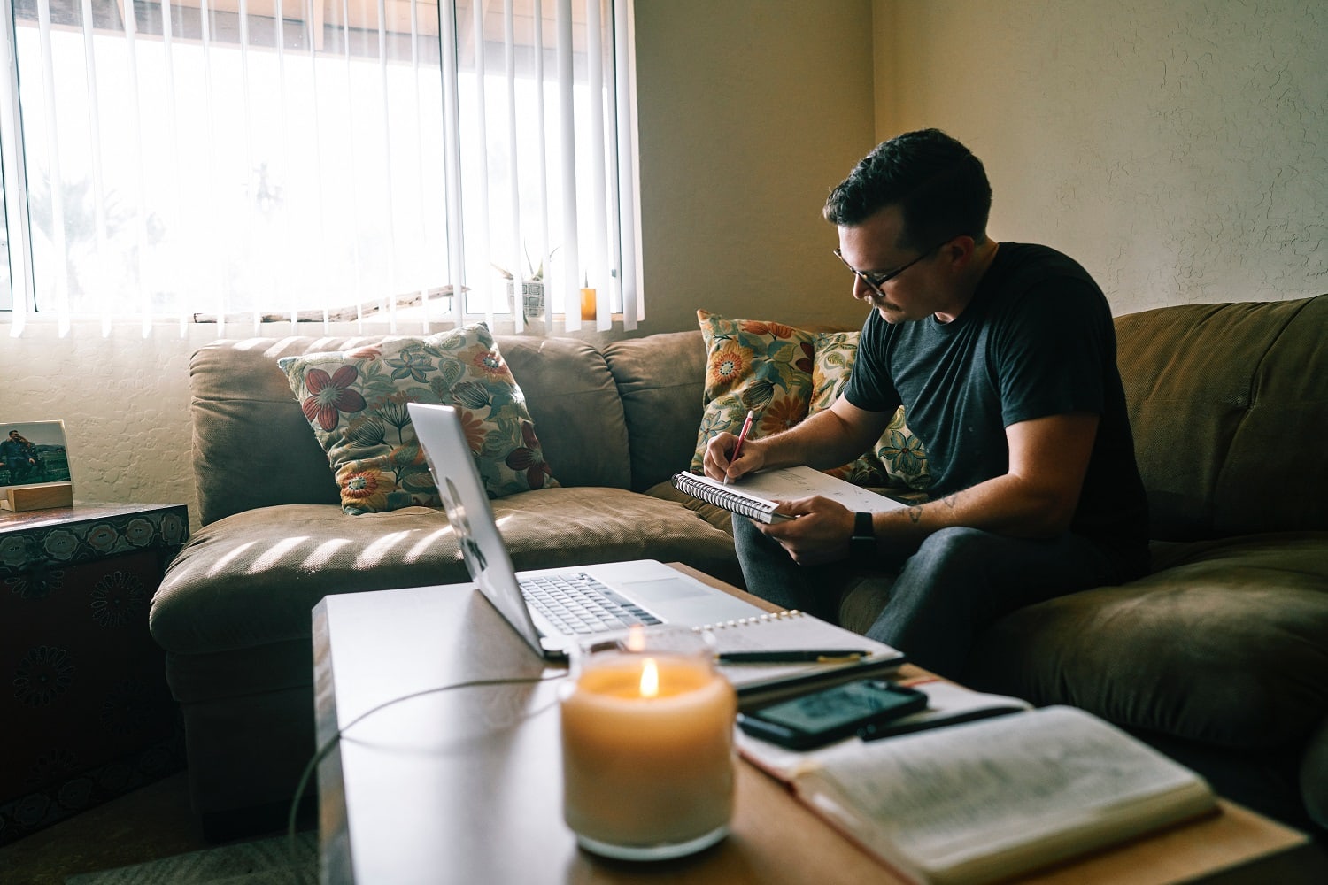 Man sits on green living couch while researching loan forbearance with a laptop and notepad. Most loan servicers recoup loan payments at the end of forbearance periods.
