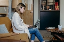 a young woamn wearing a sweater, jeans, and glasses, sits on her couch and looks at a laptop for updates on HELOCs