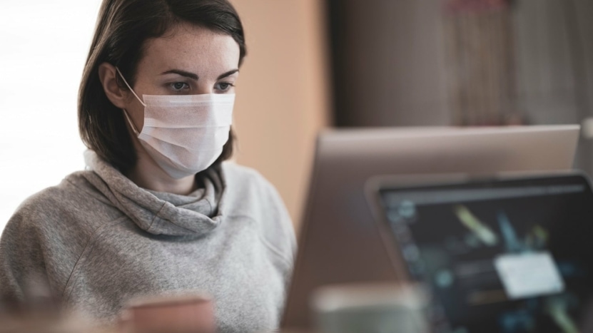 A woman wearing a respiratory mask is using her laptop at home to look up her credit score and check if it has been impacted by her mortgage forbearance.
