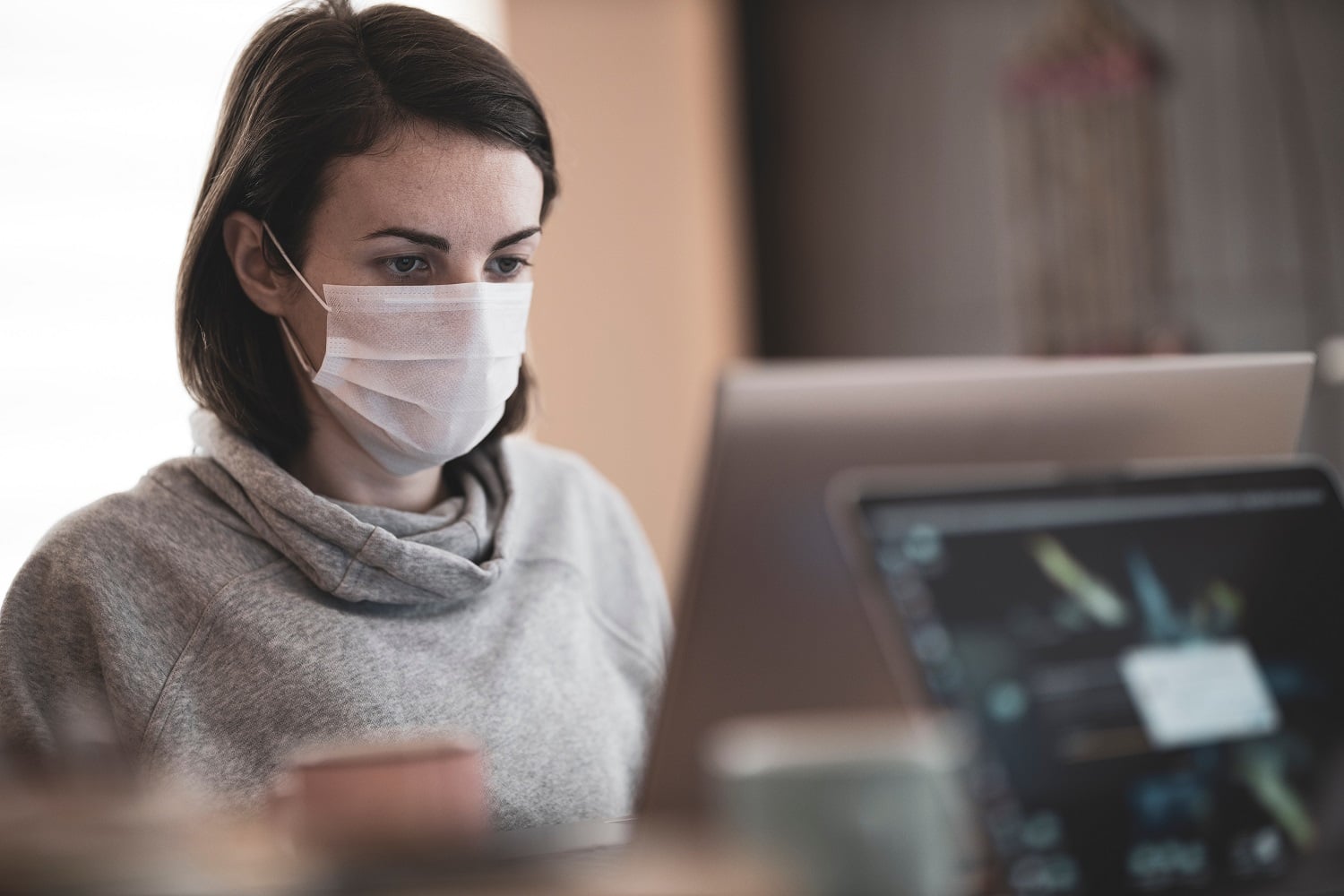 A woman wearing a respiratory mask is using her laptop at home to look up her credit score and check if it has been impacted by her mortgage forbearance.