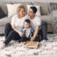 A family of three sit on the floor on a white shag rug in front of a tan couch eating pizza together