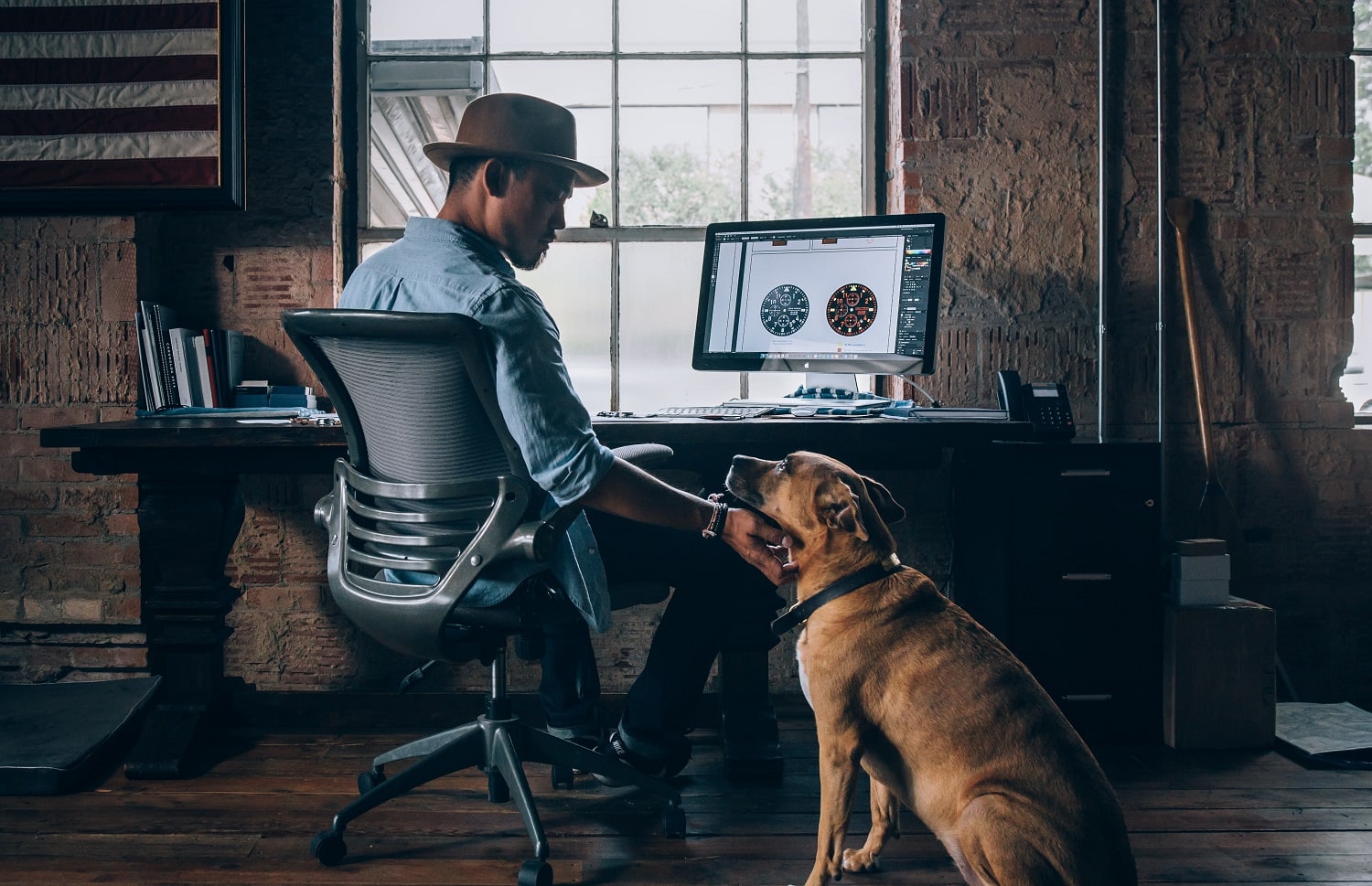 a self employed man wearing a hat sits at his desk and pets his large brown dog