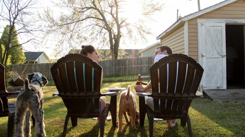 a couple sit in their backyard with dinner and drinks as they enjoy dinner with their two dogs