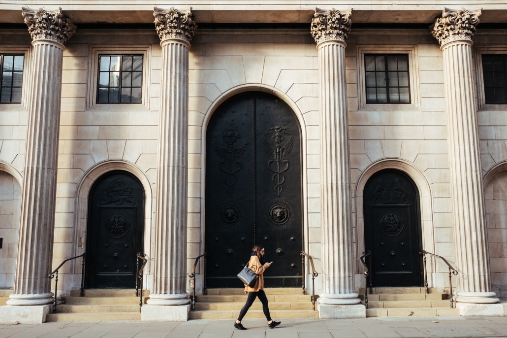 A woman walking past a large and old bank made of white stone columns. She is wearing a brown coat and is looking at her phone while reading about the differences between a non-bank and a broker for getting a mortgage.