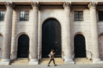 woman walking past a large and old bank made of white stone. She is wearing a brown coat and is looking at her phone while reading about the differences between a non-bank and a broker for getting a mortgage