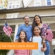 A young veteran family smiles in front of their new home with American flags. Using a VA home loan is one of the best options for veteran buyers.
