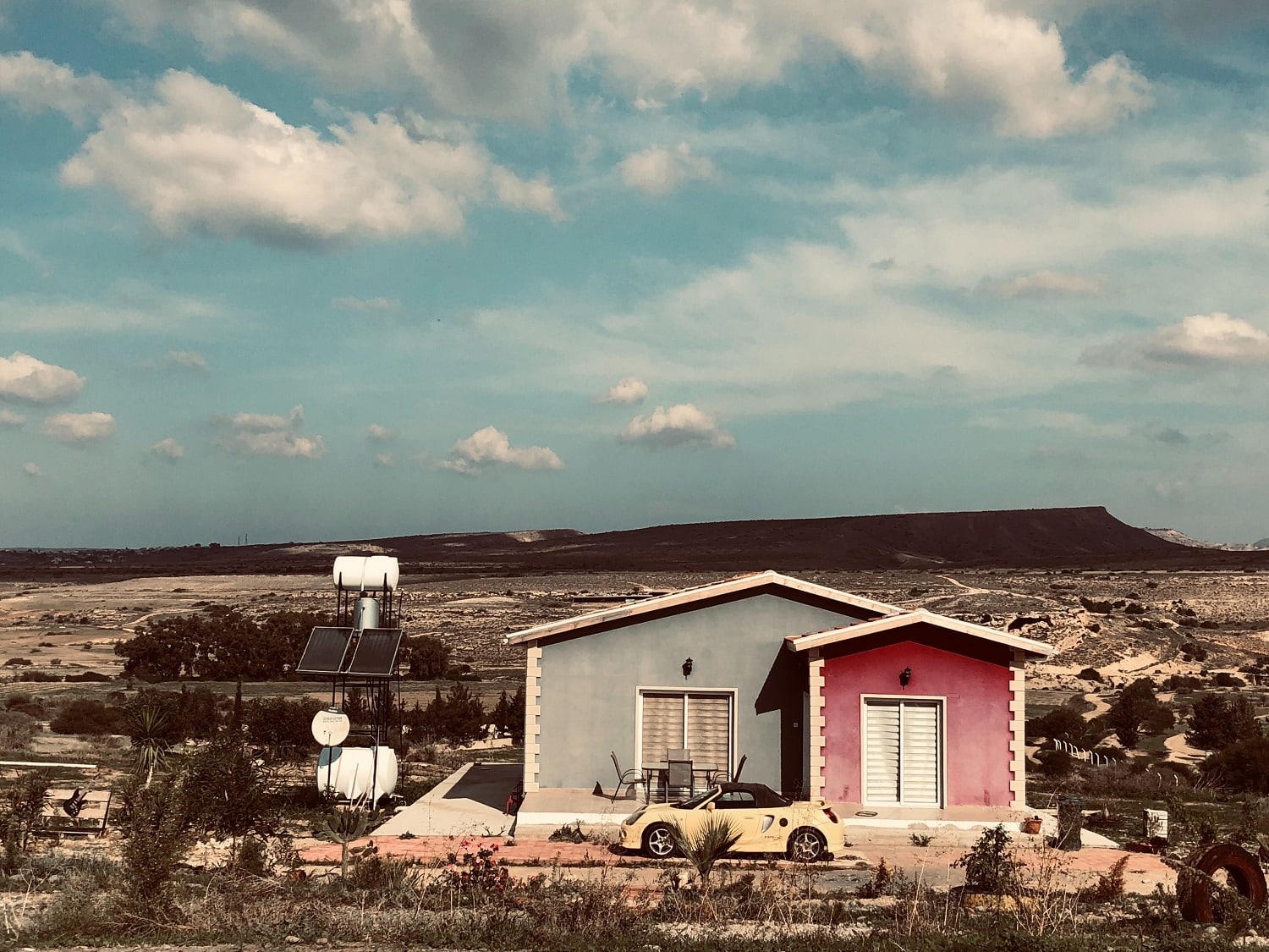 A yellow car is parked in front of a red and blue house in the desert.. Cars are items that fall under consumer price inflation but real estate is an asset and is a great hedge against inflation.