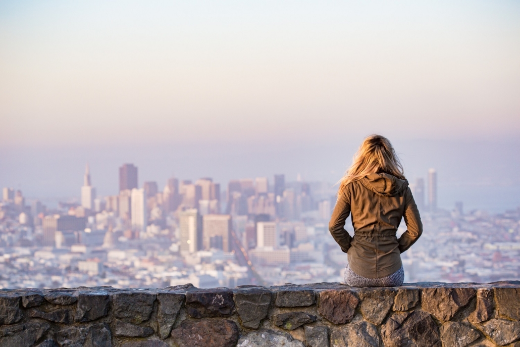 woman wearing a green jacket sitting on a stone bench with her back to the camera, overlooks San Francisco city skyscrapers with large offices