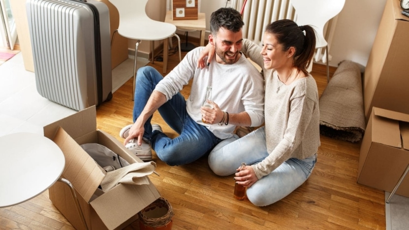 A young couple relaxes with a cool beverage while sitting in a room full of boxes. They are packing for their move after being pre-approved for a new purchase they bought with the gift of equity from a relative.