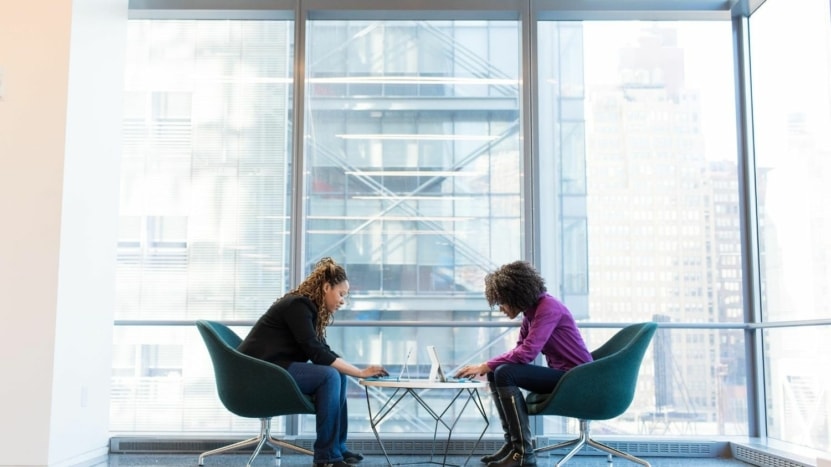 Two women sit at a table working on their laptops