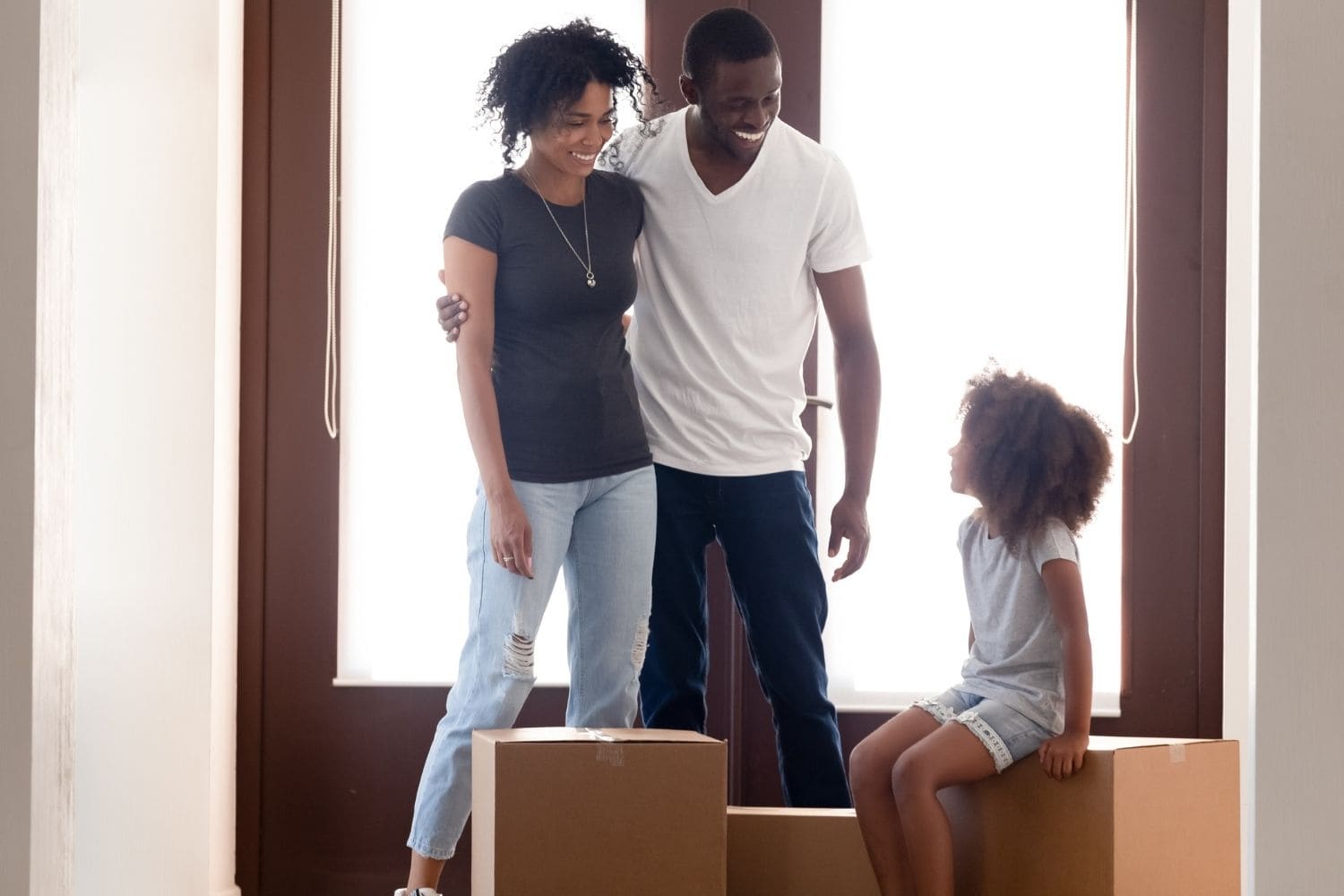 a family of three stand in the doorway of their new home the mom and dad stand with their arms around one another while their curly haired daughter sitting on a moving box