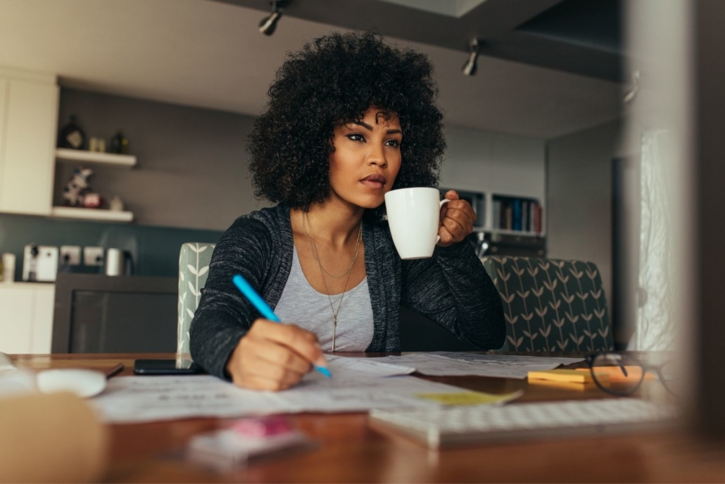 woman sits at a table in her home holding a cup. She is looking at a computer screen and holds a pen in her other hand as she works to complete her closing paperwork in order to fund the new home loan