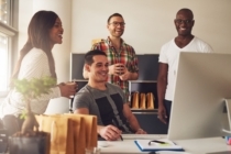 three young entrepreneur friends gather around a colleague sitting at a desk looking at a computer screen