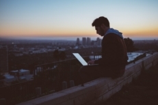 A young man sits on a rooftop after returning to work from a COVID layoff and uses a laptop.