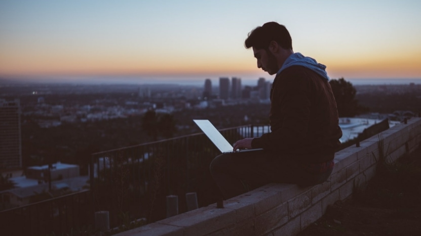 A young man sits on a rooftop after returning to work from a COVID layoff and uses a laptop.
