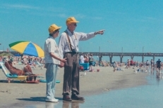 An elderly couple at the beach wear vintage clothes and look out with nostalgia and reminisce about the good old days.