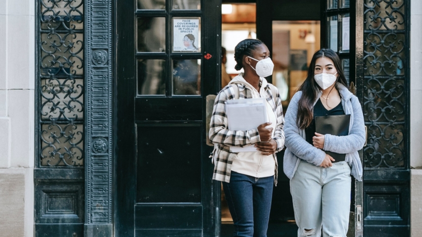 Two women wear respiratory masks to protect them from COVID stand in front of a residential real estate building with mortgage paperwork in hand.