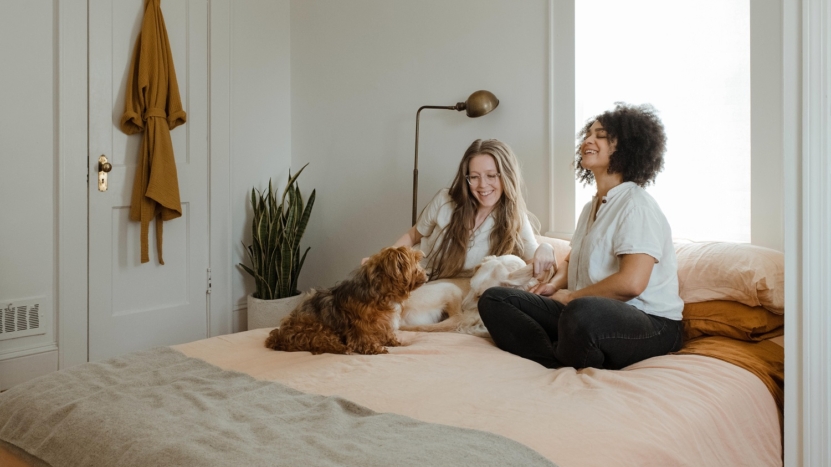 Two young women sit on the bed in their new home that they just got their offer accepted on after using these 8 tips.