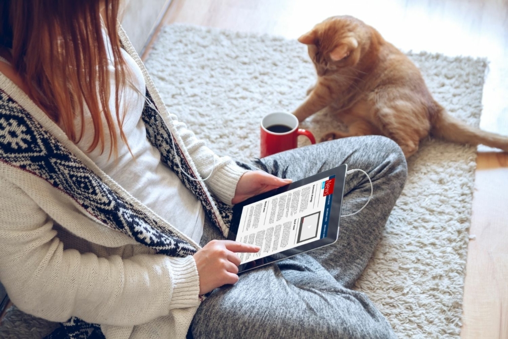 Woman sits on floor reading news on an iPad about the mortgage interest rates climb. An orange cat sits on the floor next to her.