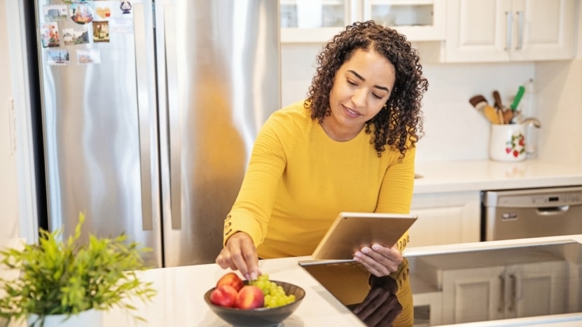 Woman eating fruit in her kitchen looking at tablet