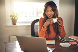 A woman wearing an orange top looks at a laptop computer while holding a cup of coffee and reviews her pre-approval letter.