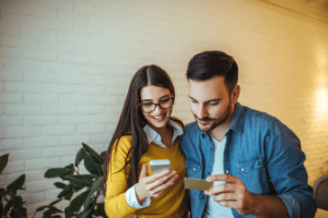 A woman in a yellow shirt and a man in a blue shirt look at a phone and credit card to review their down payment savings.