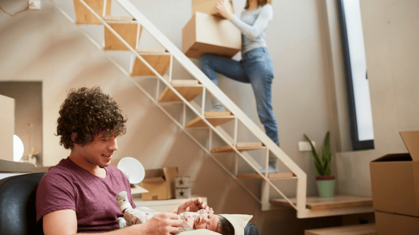 Man holds baby on the couch while woman carries moving box up the stairs