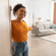 Cheerful African American Woman Opening Door And Gesturing Welcoming You To Come In Smiling To Camera Standing At Home. Hospitality, Real Estate Ownership And Purchase Concept