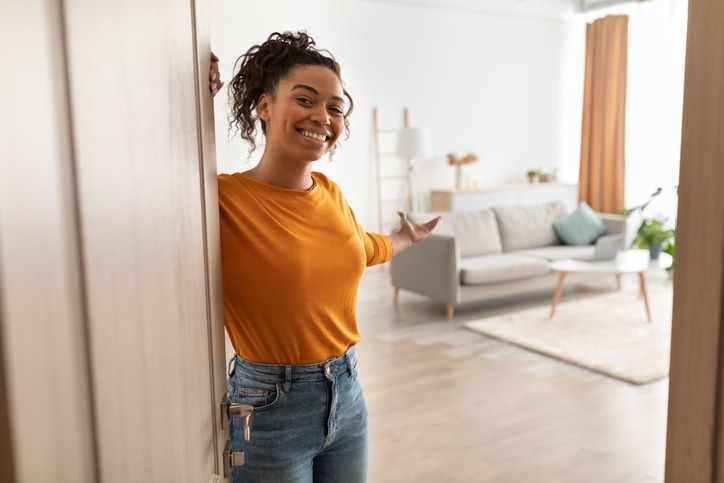 Cheerful African American Woman Opening Door And Gesturing Welcoming You To Come In Smiling To Camera Standing At Home. Hospitality, Real Estate Ownership And Purchase Concept