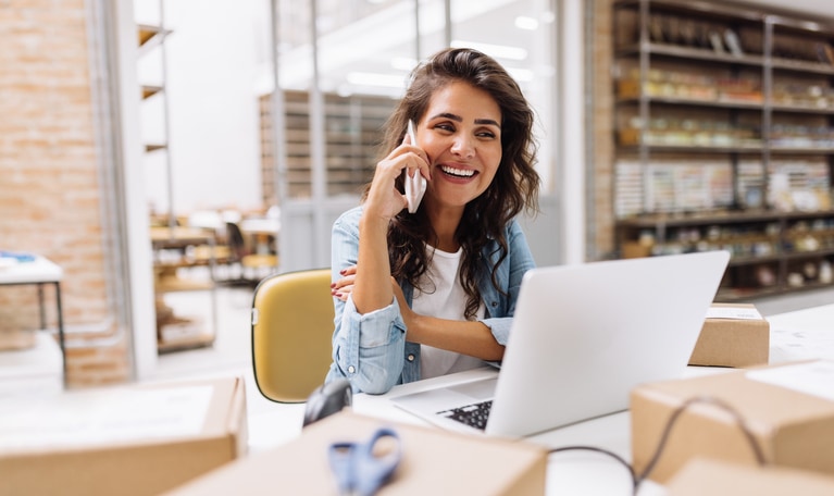Happy young businesswoman speaking on the phone to JVM Lending about a bank statement loan while working in a warehouse. Online store owner making plans for product shipping. Creative female entrepreneur running an e-commerce small business.