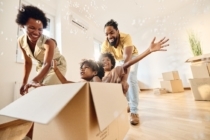 Playful African American parents having fun while pushing their small kids in carboard box at new apartment. Copy space.