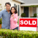 Hispanic couple outside home with sold sign holding keys in hand looking at camera smiling