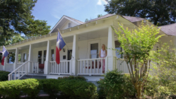 Historical home with Texas flags adorning the posts on the front porch with woman standing next to them.