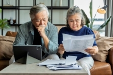 Older retired Asian senior couple checking and calculate financial billing together on sofa involved in financial paperwork