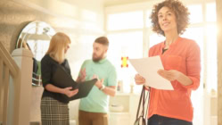 A potential homebuyer holding a property fact sheet and looking around a house with their spouse talking to a realtor in the background.