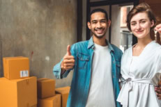 Two first-time homebuyers standing in front of moving boxes with thumbs up