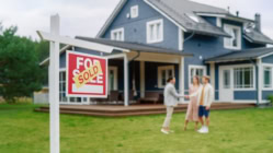 Two new homebuyers standing in front of a house with a for sale sign