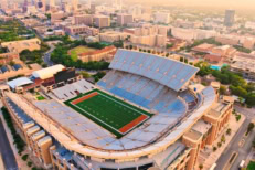 Aerial view of a football stadium in the heart of a city at sunset, with empty stands and a green field.