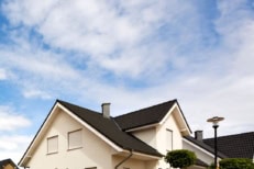 Modern suburban two-story house with dark roof tiles and cream exterior under a bright blue sky with clouds.